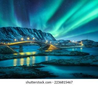Bridge and northern lights over snowy mountains. Lofoten islands, Norway. Aurora borealis and reflection in water. Winter landscape with starry sky, polar lights, rocks, road, sea, city illumination