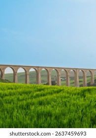 A bridge over grassy fields with a cloudy blue sky above and a hill in the distance. The arches are brown brick. Location: Tunisia, Beja ,.