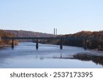 a bridge spanning a calm river with reflections on the water. The bridge is set against a backdrop of forested hills displaying autumn colors, and in the distance, two tall smokestacks rise above the 