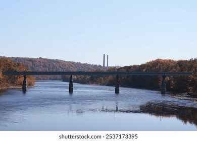 a bridge spanning a calm river with reflections on the water. The bridge is set against a backdrop of forested hills displaying autumn colors, and in the distance, two tall smokestacks rise above the  - Powered by Shutterstock