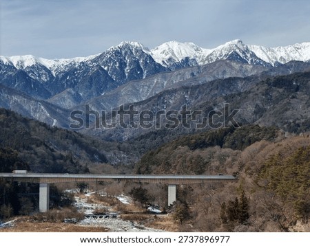 A bridge spans a river in front of a mountain range. The bridge is a long, thin structure that is suspended over the water. The mountains in the background are covered in snow
