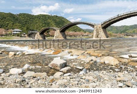A bridge spans a river with a rocky shoreline. The bridge is surrounded by trees and a small village