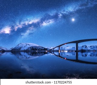 Bridge and starry sky with Milky Way over snow covered mountains reflected in water. Night landscape with road, snowy rocks, sky with moon, milky way, stars, sea. Winter in Lofoten islands, Norway 