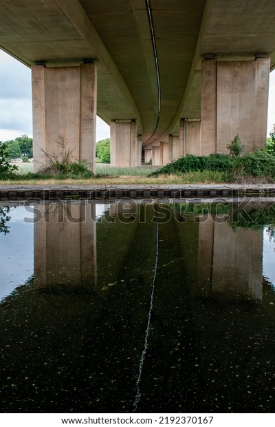 A bridge from underneath reflected in water. A\
dividing line between the two carriageways creates a stark white\
line meandering as a reflection across the dark black water.\
Strongly contrasting image.