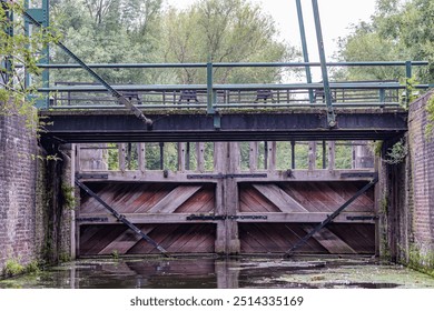 Bridge at wooden gate at entrance or exit of manual lock number 19, small boat navigation canal outside city of Maastricht, foliage of trees in background, cloudy day in South Limburg, Netherlands - Powered by Shutterstock