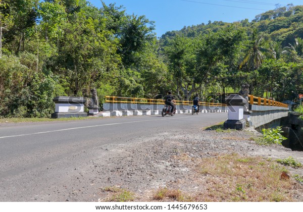 Bridge with yellow metal railing over river in\
mountains on Bali island. Decorative stone statues. People ride\
motorbikes. Passengers on the means of transport. In the background\
tropical vegetation a
