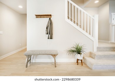 Bright and airy entry foyer with white wall stair case light colored hard wood flooring dark walnut front door entry coat rack hooks to a welcoming interior - Shutterstock ID 2298680339