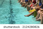 A bright aqua blue swimming pool with students sitting dangling their feet and toes in the water spectating. High school swimming carnival or club race meeting. 