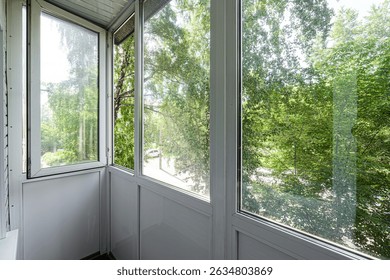Bright balcony with white frames and large windows overlooking lush green trees, bringing the outdoors in. One window is slightly open.