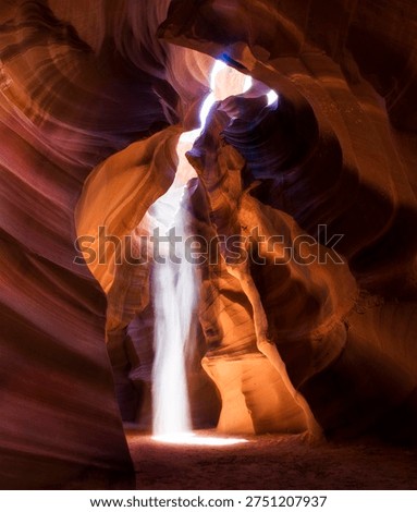 A bright beam of sunlight streams through an opening in a slot canyon, creating a bright vertical shaft of light against the curved sandstone walls. The rock displays layered erosion patterns in shades of orange, purple, and red.