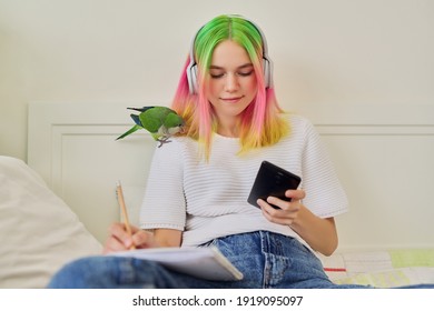 Bright Beautiful Owner Of Parrot Teenager Student With Pet On Her Shoulder. Trendy Dyed Hair Girl With Headphones Writing In A School Notebook, Using Smartphone To Study