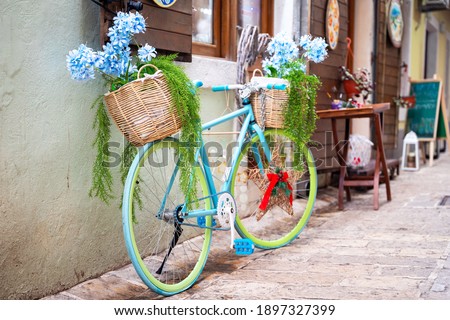 Bright bicycle decorated with baskets of pots with beautiful artificial blue flowers and dense foliage adorns the entrance to souvenir shop or authentic cafe in old town.