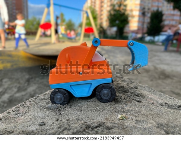 Bright blue and\
orange toy excavator with big wheels stuck in sand. A kid abandoned\
their toy car on a\
playground