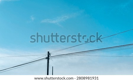 Bright blue sky with light clouds above power lines. Tall pole holds multiple cables stretching across frame. Daylight scene shows urban infrastructure and open atmosphere. Background provides clear space.