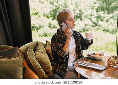 In a bright cafe, a stylish non binary person enjoys a warm drink while chatting on the phone.