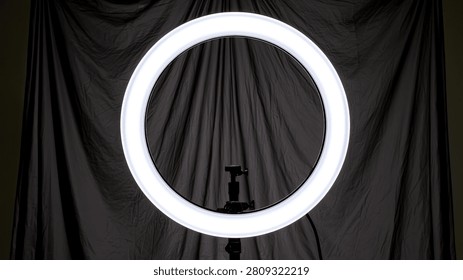 A bright, circular ring light is centered against a dark, draped fabric background, creating a dramatic and focused lighting effect.