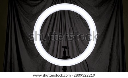 A bright, circular ring light is centered against a dark, draped fabric background, creating a dramatic and focused lighting effect.