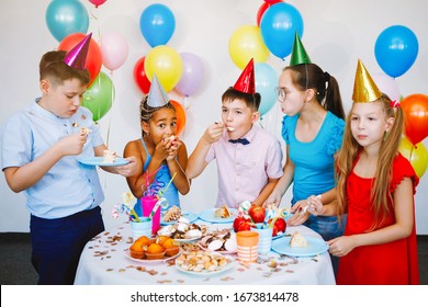 Bright, Cute Children Celebrate A Birthday. Multinational Party, Balloons, Caps, Smiles, Teens Near The Table With A Cake.