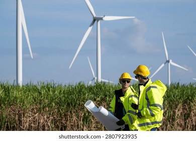 In bright diffused daylight, two turbine engineers with rolled drawings gesture over a composite blade lying in the grass at a blade laydown area in deep grass. Clean energy production.