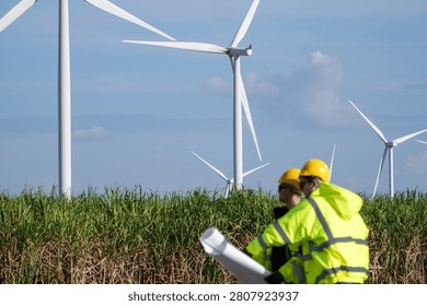 In bright diffused daylight, two turbine engineers with rolled drawings compare the blade laydown area against the plan at a blade laydown area in deep grass. Renewable electricity, green.