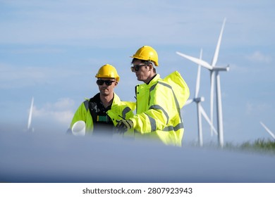 In bright diffused daylight, two turbine engineers with rolled drawings gesture over a composite blade lying in the grass at a blade laydown area in deep grass. Renewable electricity, green.