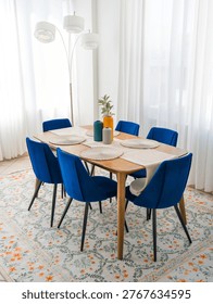 Bright dining room setup features a wooden table with blue velvet chairs next to a tall floor lamp and a large bright window with curtains. A colorful floral rug covers the tile floor in the bright apartment. Ribbed vases with green leaves and round placemats sit on the wooden board.
