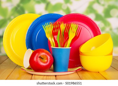 Bright disposable paper plates, cups and plastic bowls, forks, apple on an abstract green background.