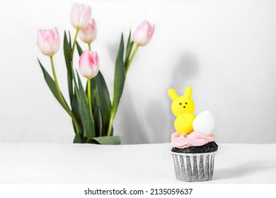 A Bright Easter Holiday, The Cupcake Is Decorated With The Favorite Marshmallow Peeps In The Shape Of A Yellow Hare And Egg. Delicate Spring Flowers Pink Tulips. White Background Of Copyspase