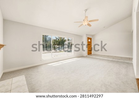 Bright, empty living room interior featuring a vaulted ceiling, wooden ceiling fan, light gray carpet, and a large front window providing ample natura