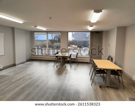 Bright, empty multipurpose room with modern laminate flooring, large windows, and tables with chairs, overlooking a patio area.