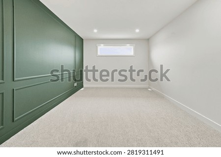 Bright, empty room features green paneled wall. White walls and light gray carpet create clean space. Small high window allows natural light entry. Recessed ceiling lights provide even illumination