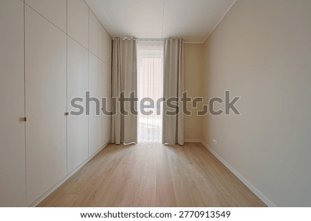 Bright empty room with full-height built-in wardrobes, sheer and blackout curtains, warm wood flooring, beige walls, and a narrow window at the end with copy space