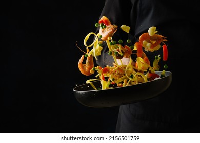 A bright festive dish - a mix of seafood, Italian pasta and vegetables in a frying pan. Explosion of colors and colors. The process of cooking seafood by a professional chef on a black background. - Powered by Shutterstock