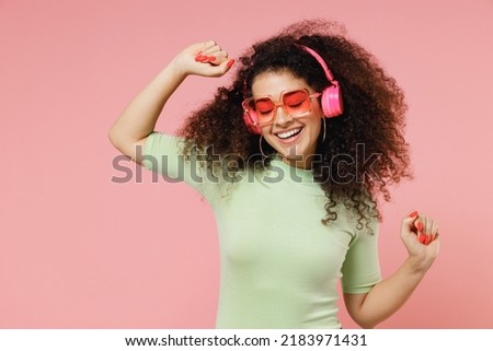 Bright fun young curly latin woman 20s wears mint t-shirt sunglasses listen music in headphones dance have fun rest relax fooling around isolated on plain pastel light pink background studio portrait