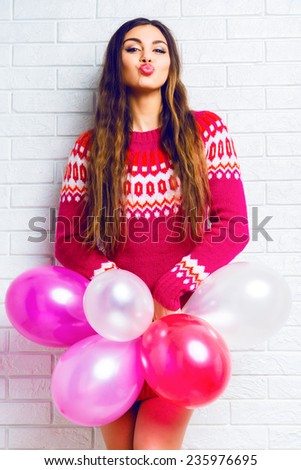 Bright funny lifestyle image to pretty young woman in casual trendy sweater having fun and holding party balloons and sending kiss. White urban background.