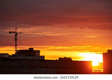 A bright golden sunset casts a warm glow over silhouetted buildings and a construction crane. The intense light filters through clouds, creating a dramatic and radiant urban scene - Powered by Shutterstock