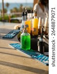 Bright green cocktail bitters bottle on bar counter in sunlight. Colorful barware and drink syrups in background with seaside blur.