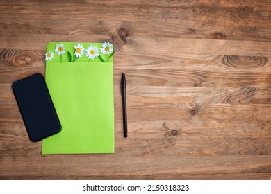 Bright Green Envelope Is Decorated With Fresh Flowers Of Daisies. The Phone And Pen Are Lying On A Wooden Textured Table. The Concept Of Flower Delivery. Online Purchase Order. 