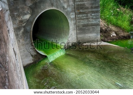 Bright green polluted effluent flowing through a drainage pipe exiting through a concrete wall in an environmental and ecological concept