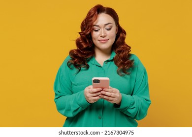 Bright happy vivid young ginger chubby overweight woman 20s wears green shirt hold in hand use mobile cell phone texting typing reading searching isolated on plain yellow background studio portrait
