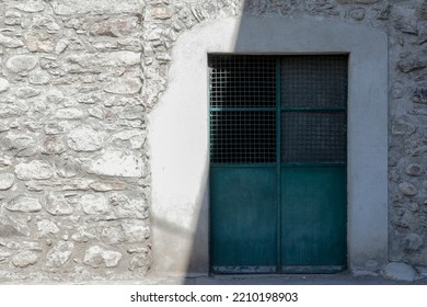 Bright House Wall With Bricks And A Green Closed Metal Door With Glass Windows, Shadow On The Door, No Person