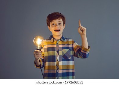 Bright Idea, Creative Solution, Discovery Concept. Portrait Of Amazed Surprised Boy Child Holding Glowing Light Bulb In Hand Gesturing Eureka Sign Pointing Finger Up Standing On Grey Studio Background