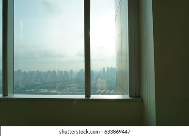 Bright interior view looking through a large modern window at a foggy city skyline with skyscrapers during morning sunrise.