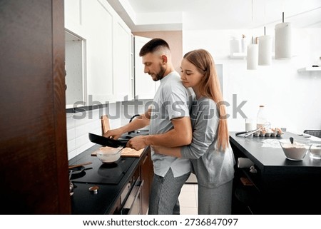 In a bright kitchen, a man is cooking while a woman stands behind him, holding him gently. They are focused on their task and sharing a moment together in the morning.
