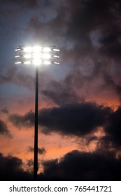 Bright LED Stadium Lights On A Tall Pole At A Sports Field, With Dramatic Orange And Blue Sunset Background And Contrasting Clouds