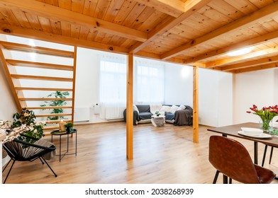 Bright Loft Apartment With Large Window, Wooden Floor And Built-in Staircase. Room Has Decorative Plants, Big Corner Sofa With Few Pillows And Nice Modern Chair With Small Coffee Table And Small Lamp.