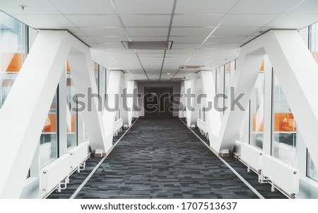 Bright long modern passageway between two towers of an office or a hospital skyscraper with  zig-zag lines of the load-bearing beams, suspended ceiling, heating batteries, and carpeting on the floor