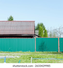 Bright metal fence and house roof in countryside. Rural house with corrugated roof behind a bright green metal fence, seen on a sunny day with blue sky and grass. - Powered by Shutterstock - Get 15% off with code: PIKWIZARD15