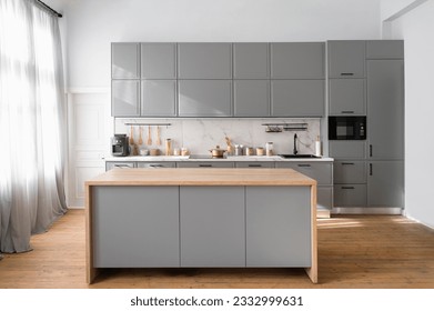 Bright minimalistic kitchen interior with gray furniture and big dining table on wooden floor. Cooking utensils and jars with food on countertop.