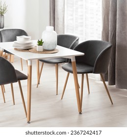 Bright Modern Dining Room with Wood-Legged White Table, Grey Fabric Chairs, White Dinnerware, Home Decor, Natural Light from a Window, and Wooden Flooring. - Powered by Shutterstock